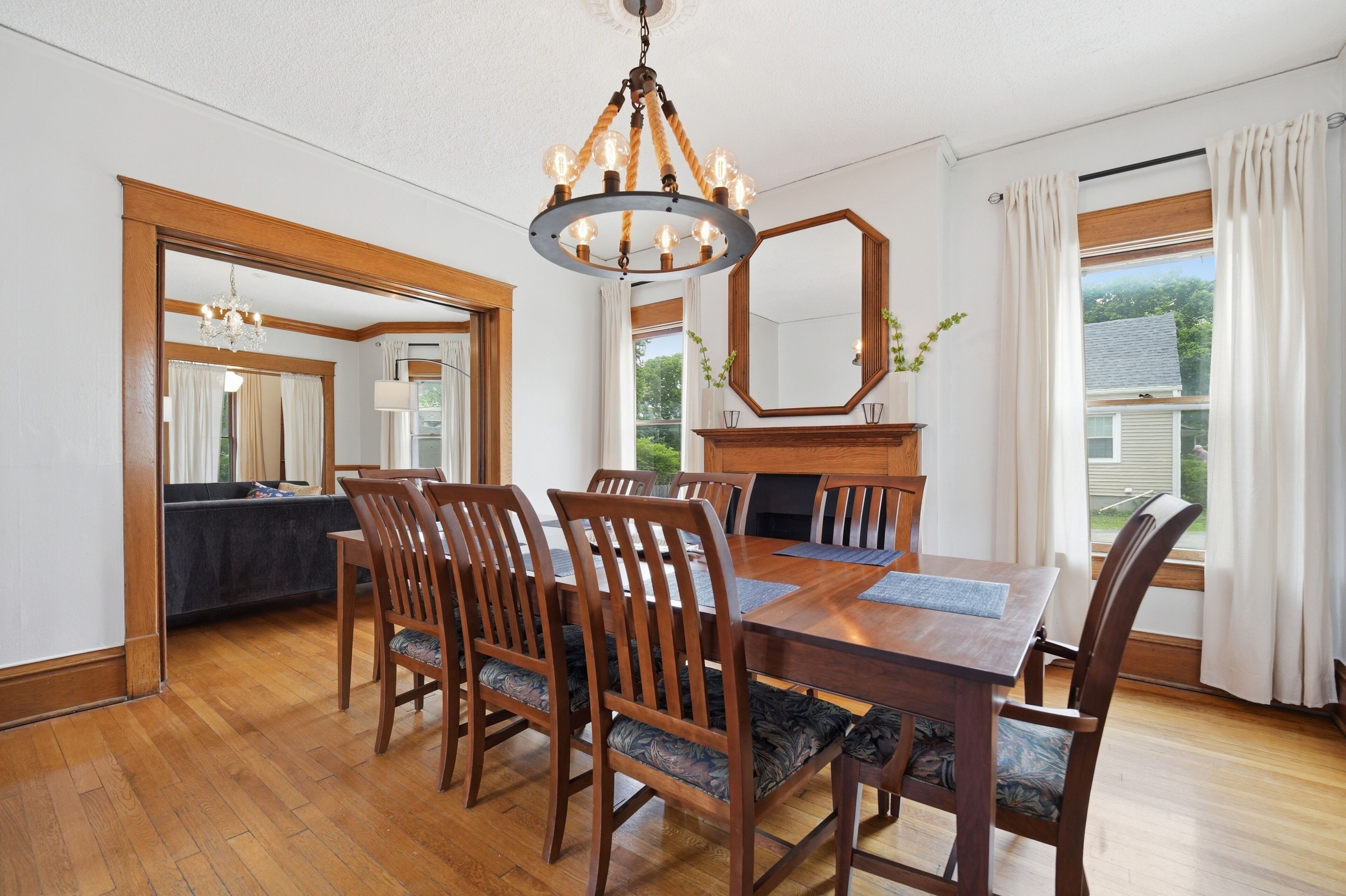 A dining room with a wooden table, chairs, a chandelier, a fireplace, and large windows with curtains.
