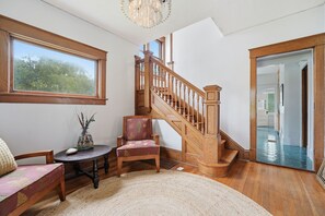 Ein Zimmer mit einer Holztreppe, einem runden Tisch mit einer Vase, einem gepolsterten Sessel und einem großen Fenster.