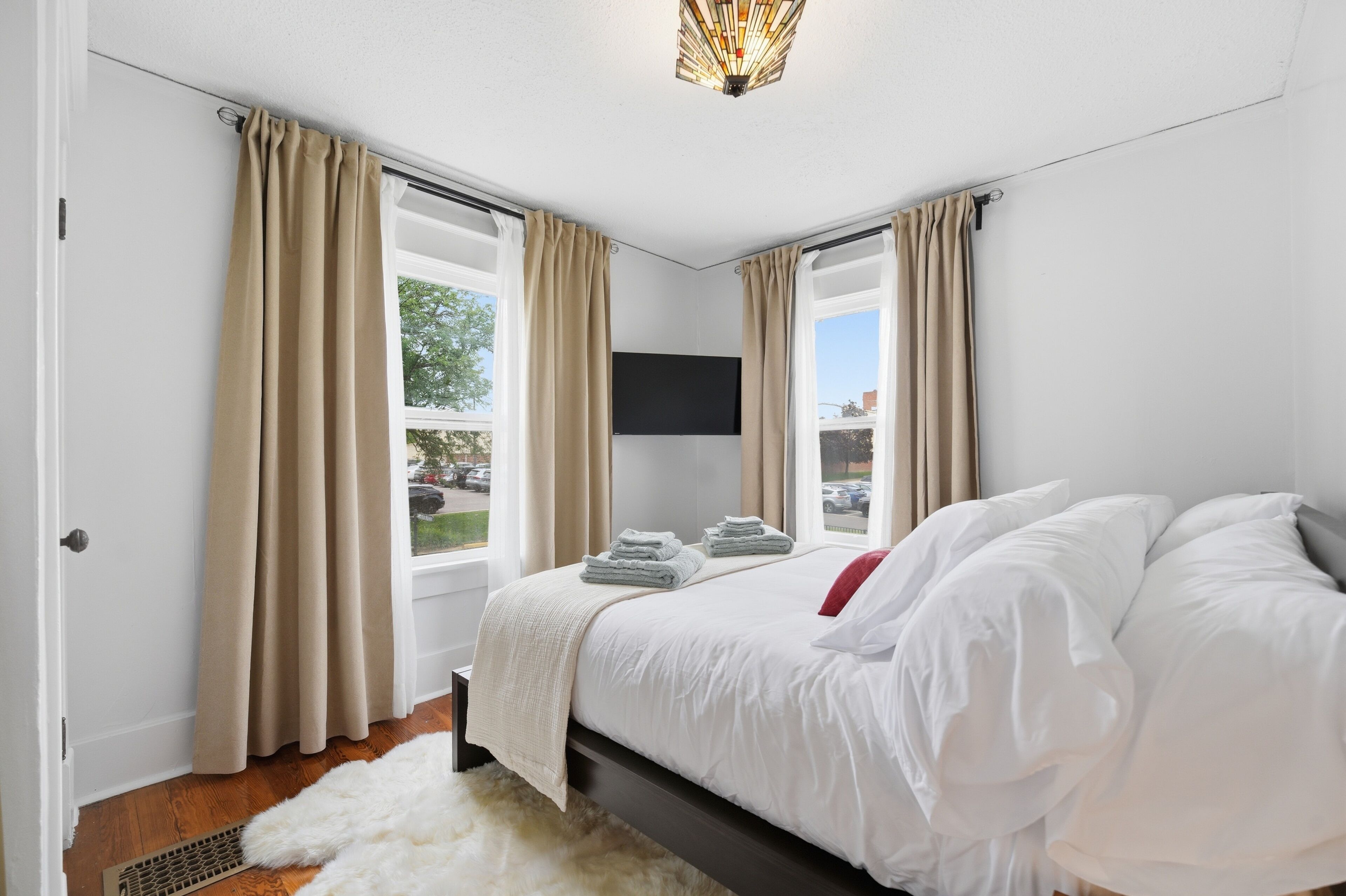 A bedroom with a bed, curtains, a flat-screen TV, and a view of the outdoors.