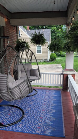 A covered porch with a hanging chair, a patterned rug, and a view of a house and greenery. - World Cup Stay! Historic KC Charmer Near KU Med & 39th Street (Kansas City)