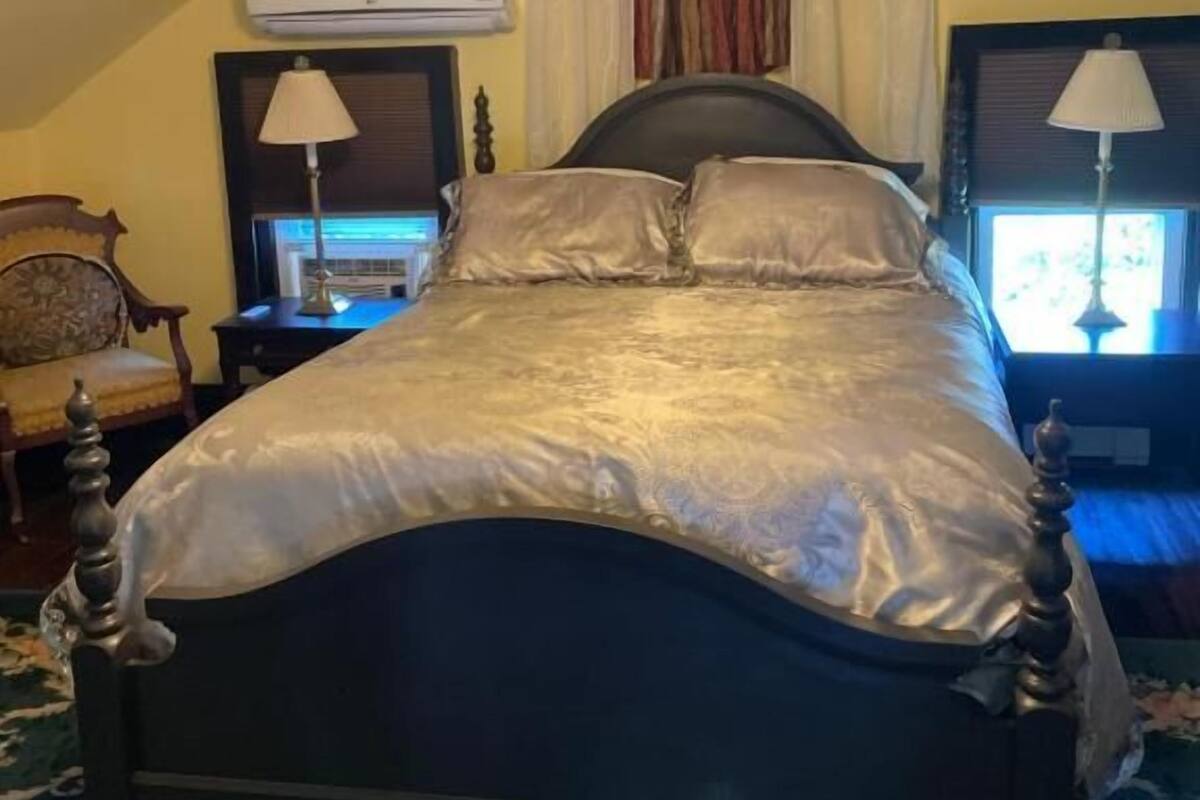 A bed with a headboard, a bedside table, a lamp, an air conditioning unit, and a window with curtains.