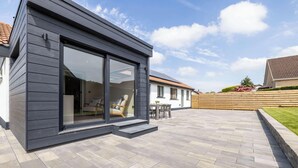 A modern house with a large sliding glass door, a patio with a table and chairs, and a well-maintained garden. - Luxury House in Templepatrick I Big Garden I Near Airport & Belfast (Templepatrick)