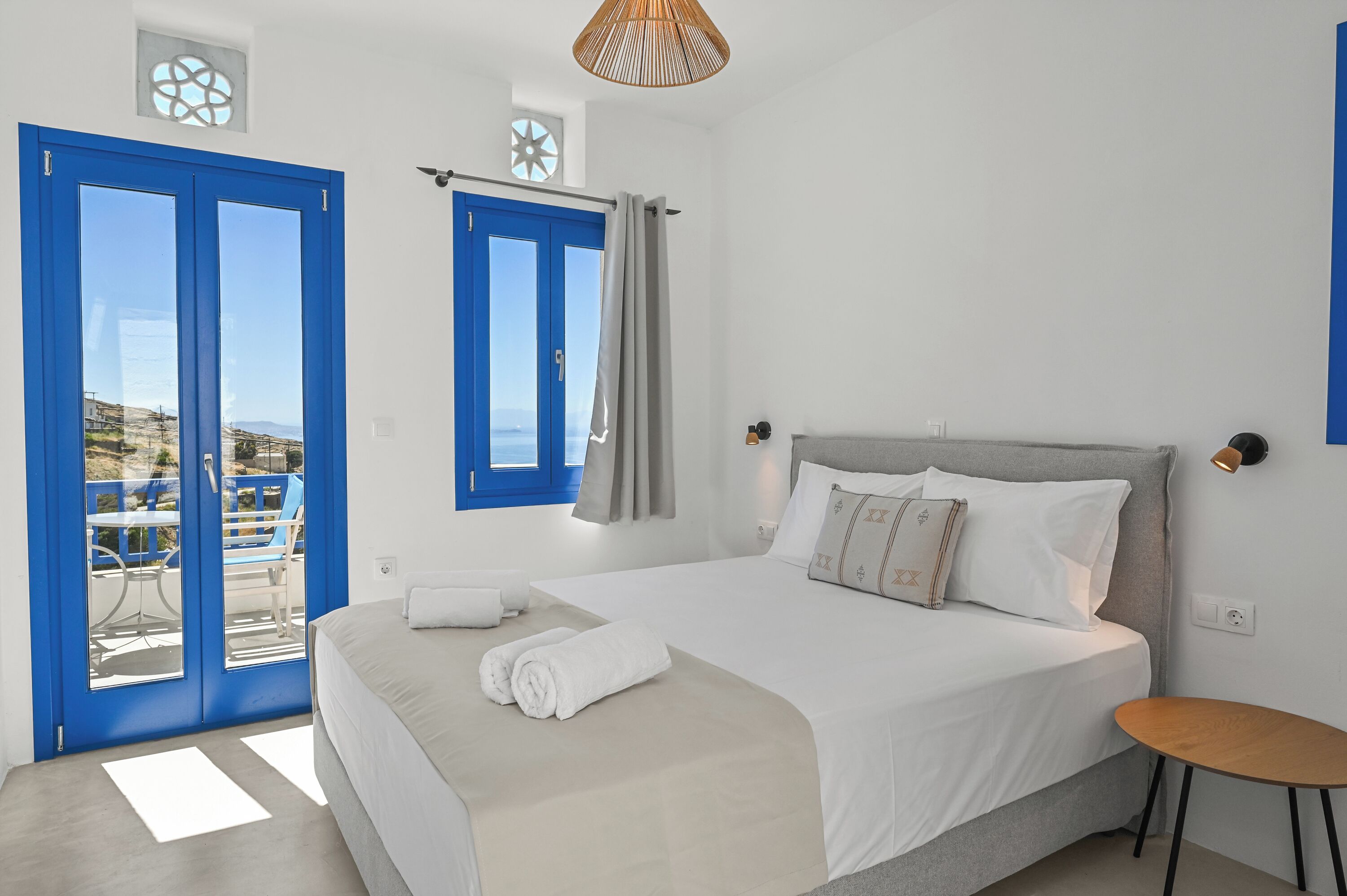 A bedroom with a bed, a small table, a window with curtains, and a view of the outdoors.
