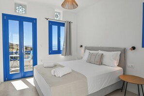 A bedroom with a bed, a small table, a window with curtains, and a view of the outdoors. - Arnados Tinos Rooms by Tinosstay (Tinos)