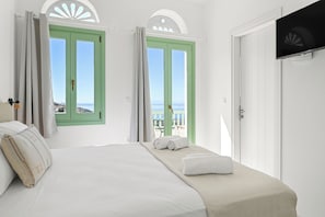 A hotel room with a bed, two green-framed windows, a TV, and a balcony with a view. - Arnados Premium Rooms (Tinos)