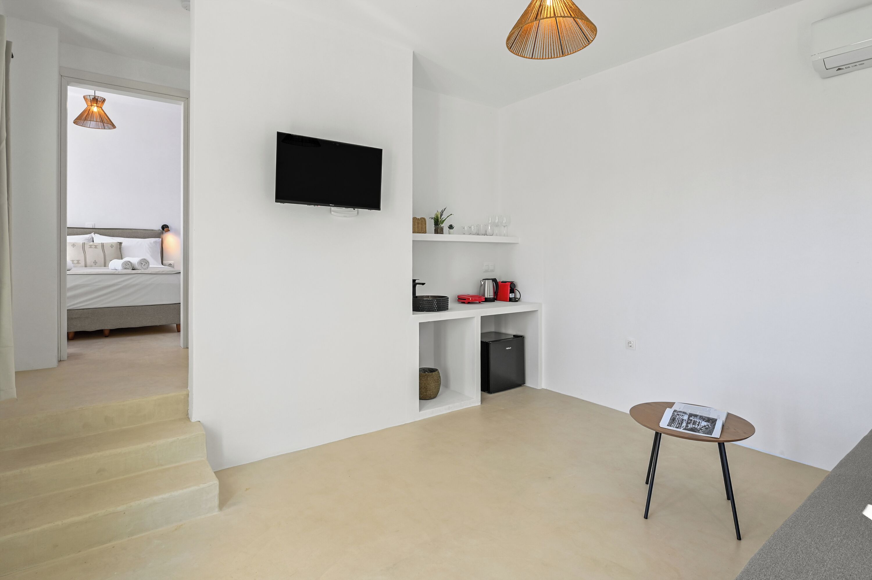 A modern, minimalistic room with a bed, a TV, a small table with a magazine, and a kitchen area with a kettle and toaster.
