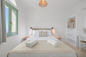 A neatly made bed with two rolled towels, a bedside table with a book, a hanging garment rack, and a window with a view. - Arnados Tinos Rooms by Tinosstay (Tinos)