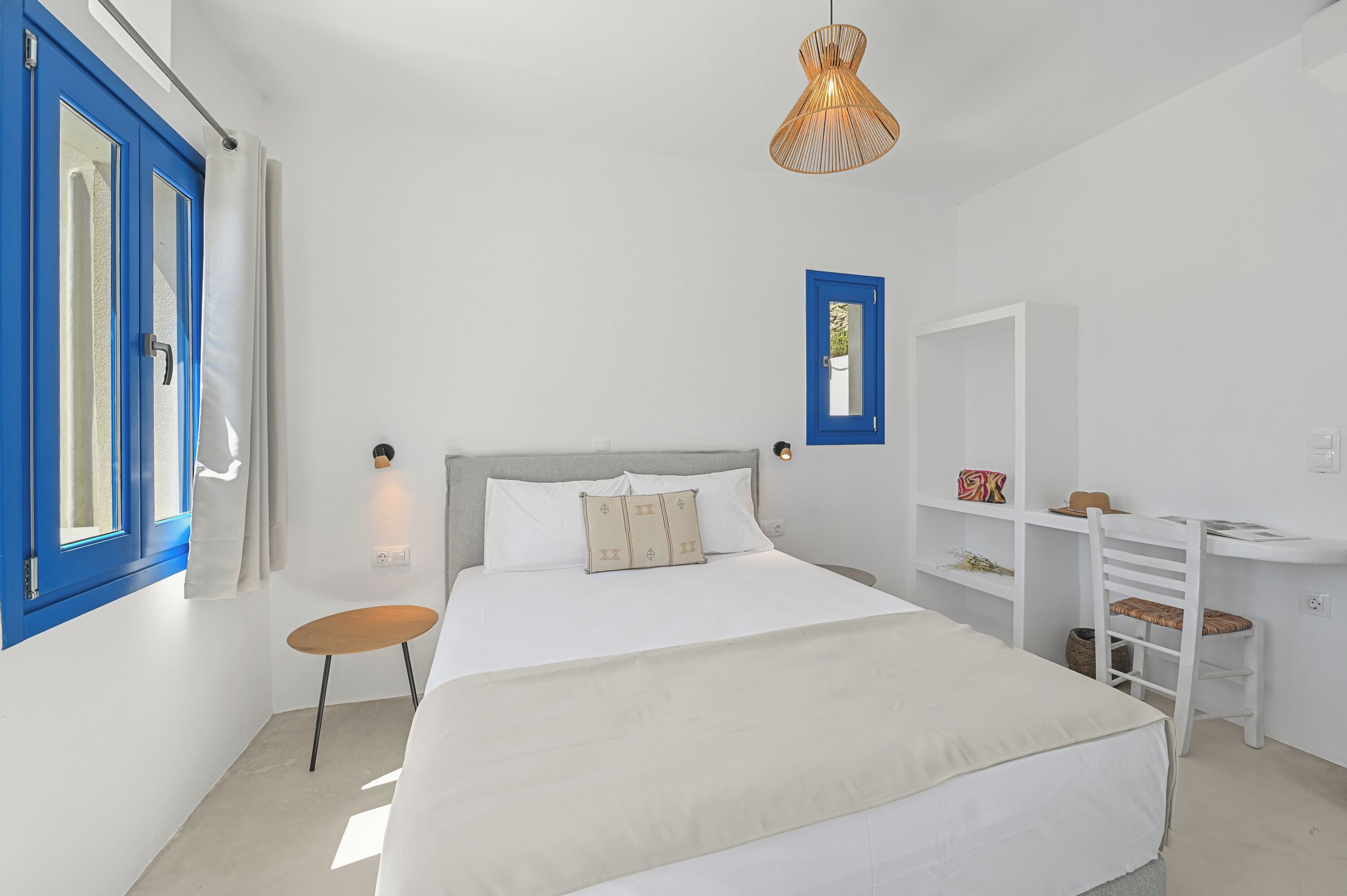 A bedroom with a bed, a small table, a chair, a desk, and a hanging light fixture.