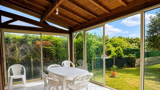 En overdækket terrasse med et bord og stole, store vinduer og udsigt over en have.