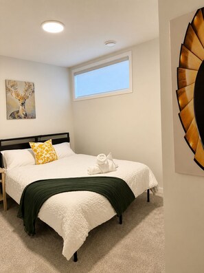 A neatly made bed with a white comforter and a green throw blanket, a yellow pillow, and a white towel on the bed. There is a painting of a deer on the wall, a window with a view, and a unique wall decoration. - 2-bedroom legal basement unit in Northwest Calgary (Calgary)
