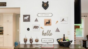 Une pièce avec un mur blanc décoré d’œuvres d’art sur le thème de la faune, une tête d’ours et une pancarte sur laquelle on peut lire « Bonjour ». On y trouve des bocaux sur le comptoir, un panier rempli d’objets et une autre pancarte qui indique « NE PAS S’APPROCHER ».