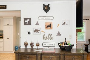 A room with a white wall decorated with wildlife-themed artwork, a bear head, and a 'hello there' sign. There are jars on the counter, a basket with items, and a sign that says 'STAY AWAY'. - Quiet North Conway home: Hot tub, arcade, outlets! (Conway)