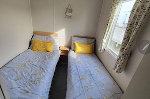 A small room with two single beds, each with a pillow that says 'this is my HAPPY place.' There is a small table between the beds, and a window with curtains.