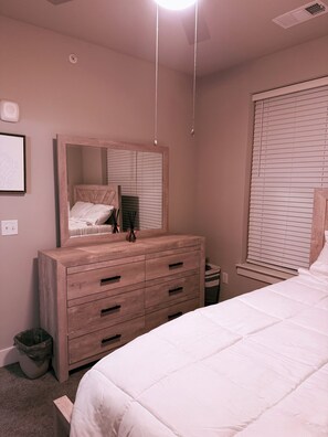 Une chambre avec une commode en bois, un lit, un miroir et une fenêtre avec des stores.