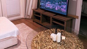 En stue med et fjernsyn på et træstel, et sofabord i glas med stearinlys, en sofa og en dør med persienner.