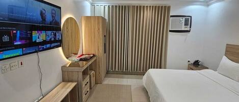 A modern hotel room with a large flat-screen TV, a wooden desk, a bed, and a ceiling light fixture.