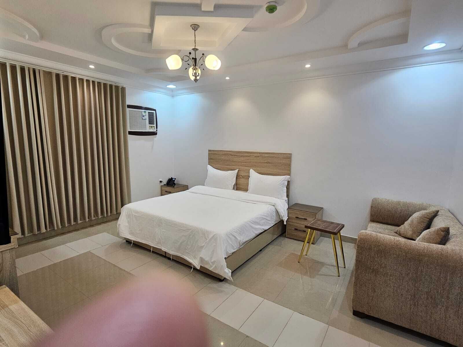 A modern hotel room with a large bed, bedside tables, a chair, a ceiling light, and an air conditioning unit.