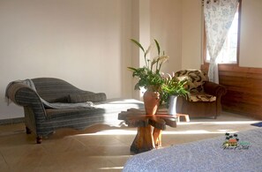 Ein Zimmer mit einem grauen Sofa, einem Couchtisch aus Holz mit einer Pflanze und einem Bett mit weißer Bettdecke.