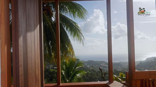 A wooden-framed window with a view of a coastal landscape, including palm trees and a distant cityscape. - Tranquil 1-bedroom apartment in charming Kingstown perfect for relaxing (Kingstown)