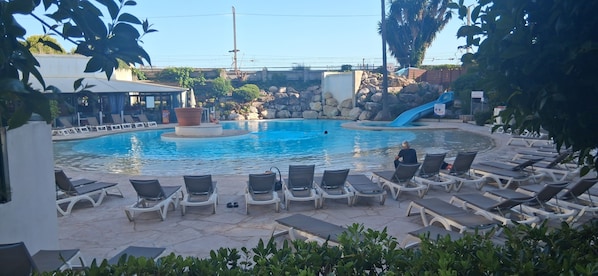 A swimming pool with lounge chairs, a water slide, and a rocky backdrop. - Garden level in residence pierre et vacances (Cannes)
