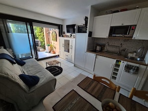 A compact living space with a sofa, a dining table, a kitchen area with cabinets and a microwave, and a sliding glass door leading to an outdoor area. - Garden level in residence pierre et vacances (Cannes)