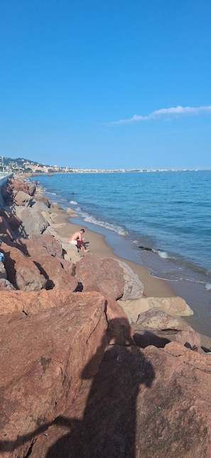 A rocky beach with a clear blue sky, a person sitting on the rocks, and a distant shoreline with buildings. - Garden level in residence pierre et vacances (Cannes)