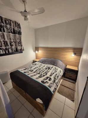 A bedroom with a bed, a ceiling fan, a wall-mounted light, a wooden headboard, and a poster on the wall. - Garden level in residence pierre et vacances (Cannes)