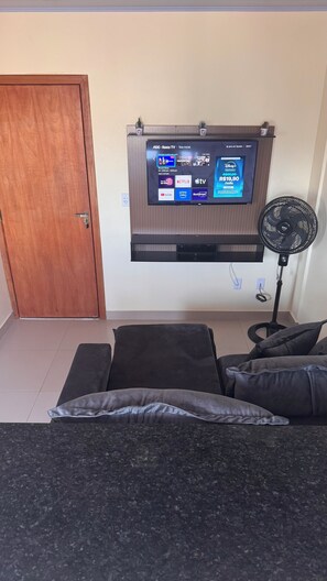Uma sala de estar com televisão na parede, um ventilador de chão e um sofá.