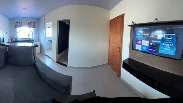 A living room with a sofa, a TV mounted on the wall, and a kitchen area in the background.