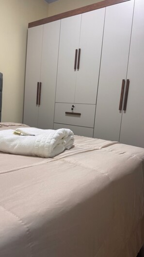 Cama com edredom branco e toalha branca dobrada. Ao fundo, guarda-roupa de madeira com duas portas e uma pequena gaveta.