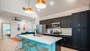 A modern kitchen with dark cabinets, a large island with stools, a microwave, and a ceiling fan. - EaDo Main House: Sleeps 12, Hot Tub Access, Near Downtown, NRG, Daikin & more (Houston)