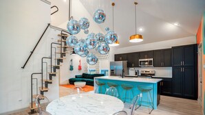 A modern kitchen with a central island, black cabinets, and a staircase with a glass railing. - EaDo Main House: Sleeps 12, Hot Tub Access, Near Downtown, NRG, Daikin & more (Houston)