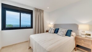 Un dormitorio con un ventanal grande, una cama con almohadas, una mesita de noche con lámpara y vistas al océano.