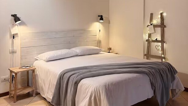 A neatly made bed with a wooden headboard, bedside lamps, and a small wooden nightstand. - Adelaide in collina, cosy home (Volta Mantovana)
