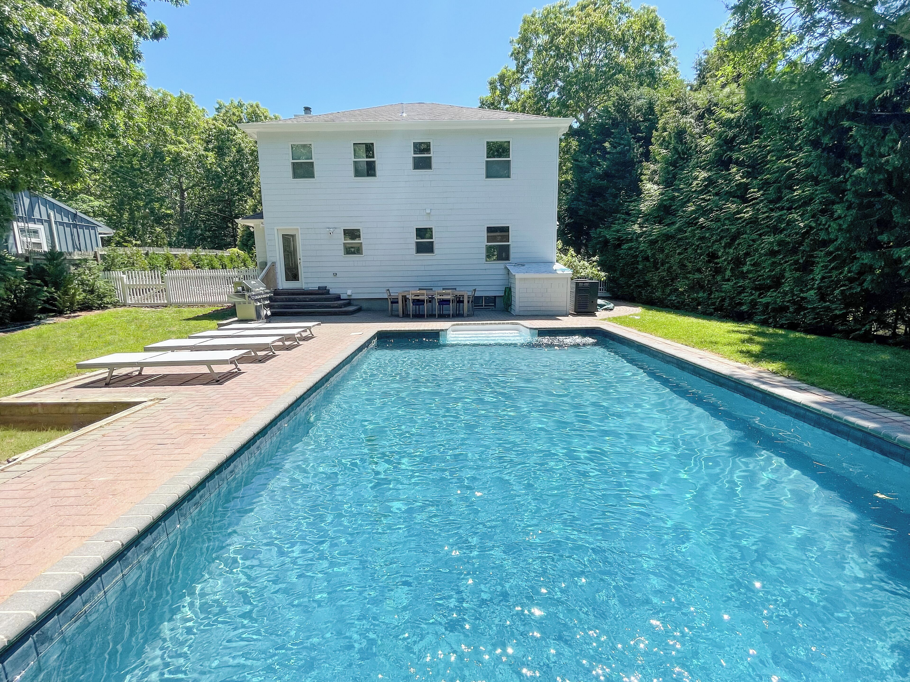 A rectangular swimming pool with a paved deck, surrounded by a grassy area and a white building with a dark roof.