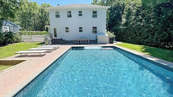 A rectangular swimming pool with a paved deck, surrounded by a grassy area and a white building with a dark roof.