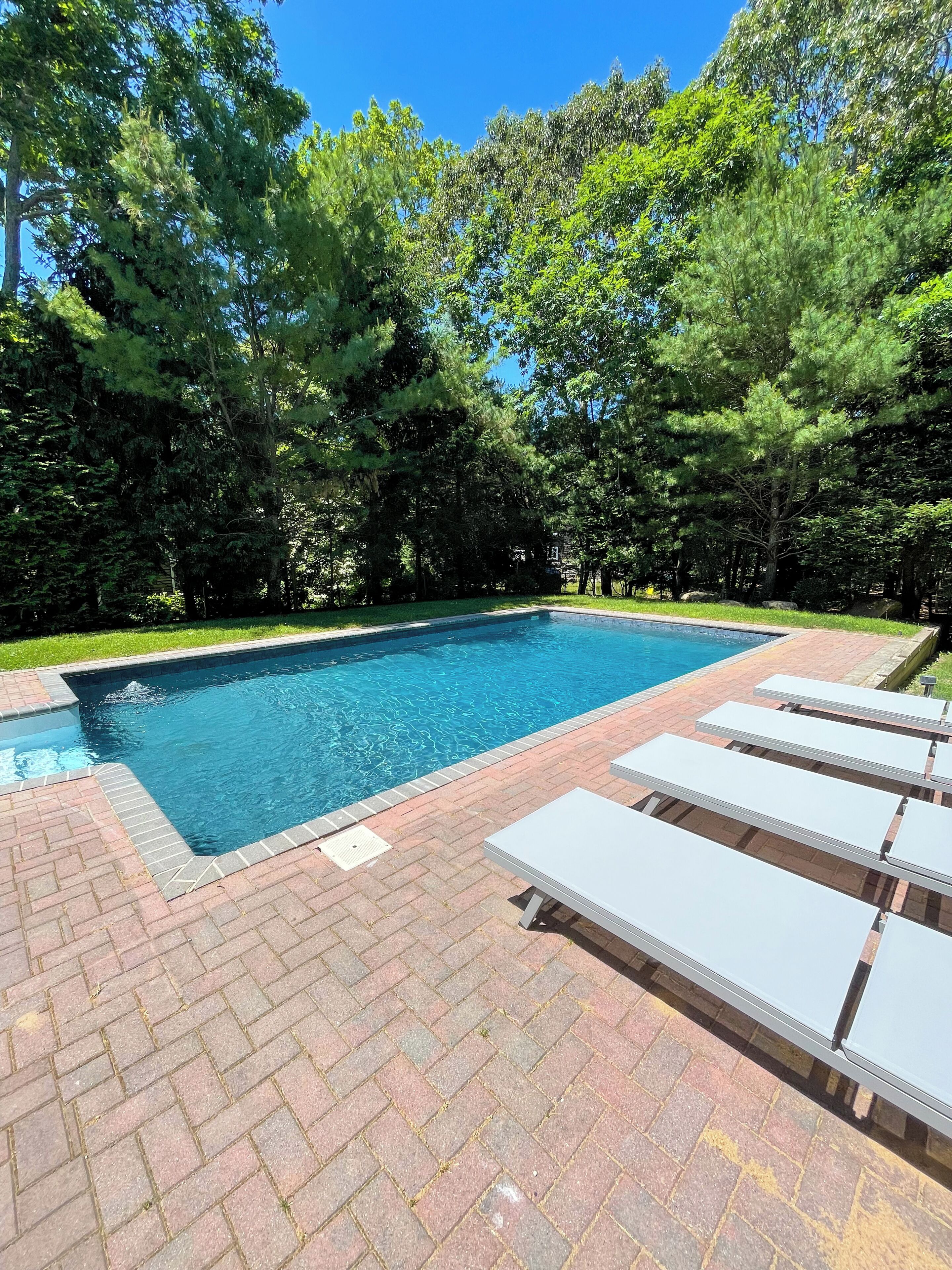 A rectangular swimming pool with a brick border, surrounded by lush green trees and a brick patio with white lounge chairs.