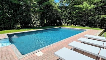 A rectangular swimming pool with a brick border, surrounded by lush green trees and a brick patio with white lounge chairs.