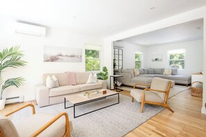 A spacious living area with a sofa, armchair, and coffee table. There is a large window providing natural light and a view of greenery outside.