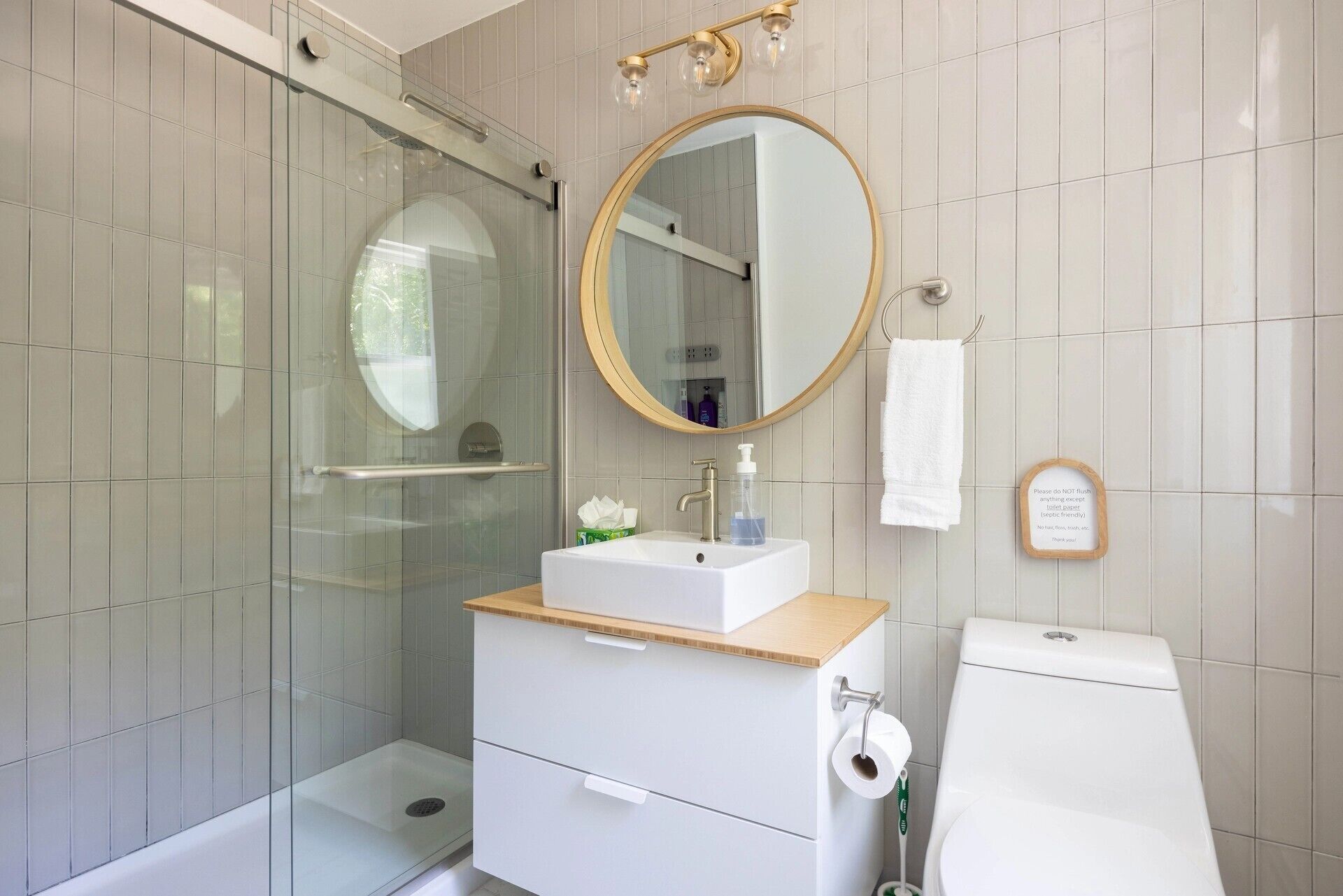 Un baño moderno con cabina de ducha de vidrio, un espejo redondo, un lavabo blanco con encimera de madera y un inodoro.