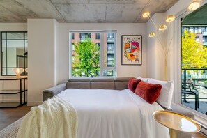 Une chambre moderne avec un grand lit, une vue sur les immeubles et les arbres, un tableau de Picasso et un balcon avec des sièges d’extérieur.