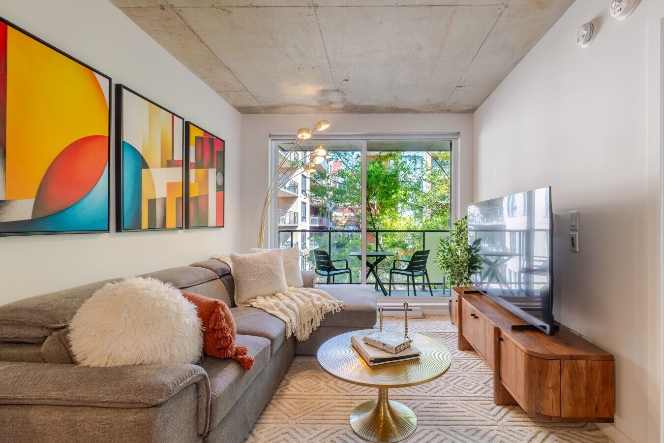 Un salon moderne avec un canapé, une table basse et un téléviseur. Les murs sont ornés de tableaux abstraits et un balcon aménagé avec du mobilier d’extérieur complète l’ensemble.
