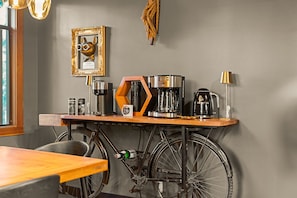 A room with a wooden table, a bicycle, a coffee machine, a framed picture, and a lamp.