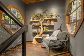 A cozy reading nook with a unique tree-shaped bookshelf, a comfortable chair, and a view through a window.