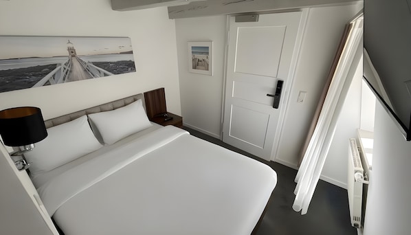 Una habitación de hotel con cama, mesita de noche, lámpara, puerta y un cuadro colgado en la pared.
