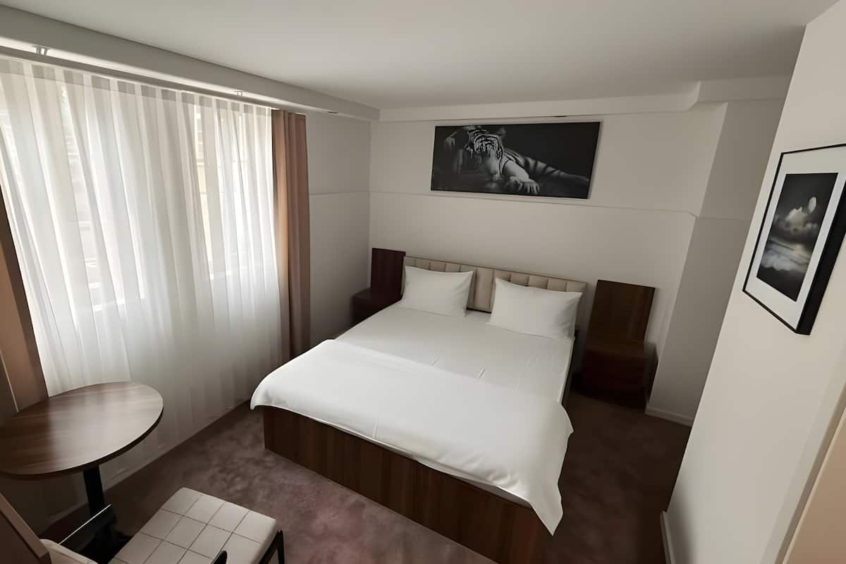 Una habitación de hotel con cama, una mesita, una silla, un ventanal con cortinas y un cuadro en la pared.