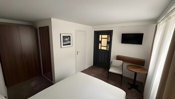 Quarto com cama, guarda-roupa, secretária com cadeira, televisão e porta com janela.