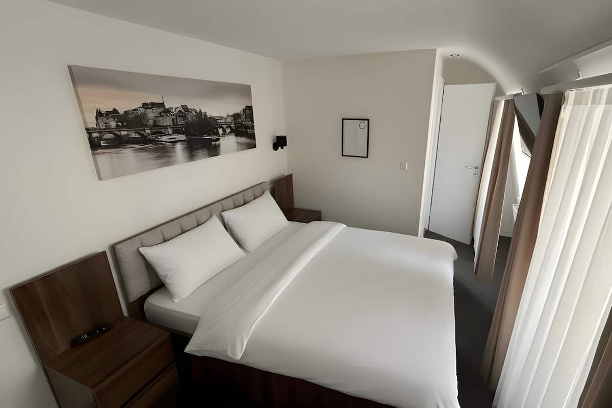Habitación de hotel con cama, mesita de noche, un cuadro, una puerta y ventana con cortinas.