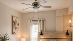 Un dormitorio con cama, mesitas de noche, cómoda, ventilador de techo, ventana con cortinas y lámpara de pared.