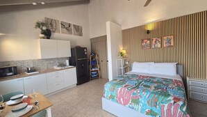 Ein kompaktes Hotelzimmer mit einer Küchenzeile, einem Bett mit floralem Überwurf, einem Deckenventilator und einer gestreiften Wand.